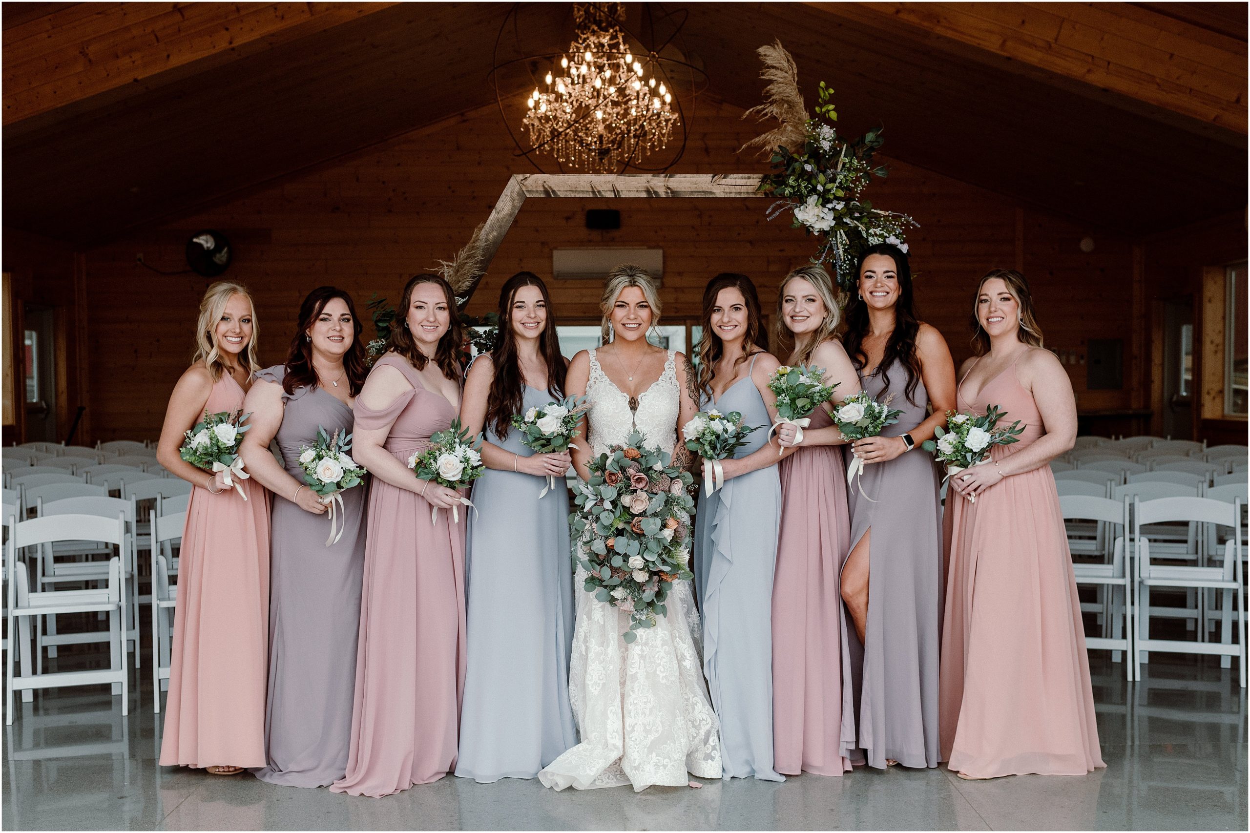 Bride and Bridesmaids smile at the camera. Bridesmaids are in spring colors of light pink, lavender, blue, and peach and holding white and green wedding bouquets at the Sparks Barn in Louisville NE by Anna Brace, who specializes in Omaha NE wedding photography.