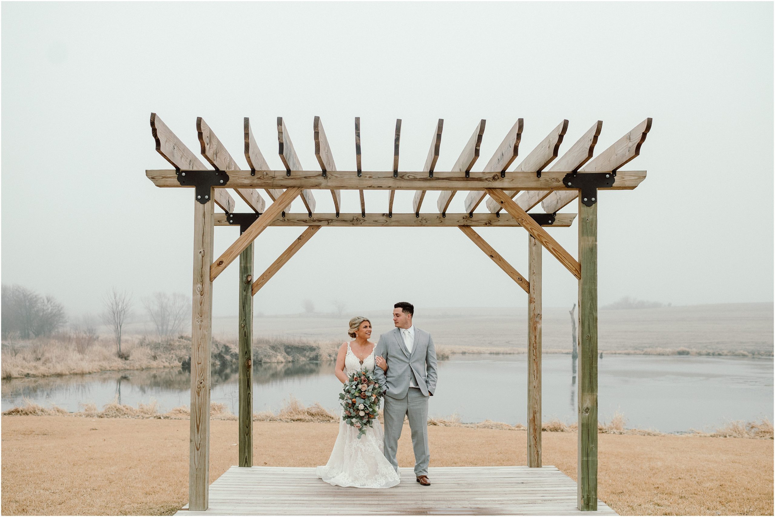 Bride in champegne and white wedding gown and groom in light gray suit with white tie stand and smile at each other underneath the pergola at Sparks Barn, a Nebraska Barn Wedding Venue. Photo by Anna Brace who specializes in Nebraska Wedding Photography.