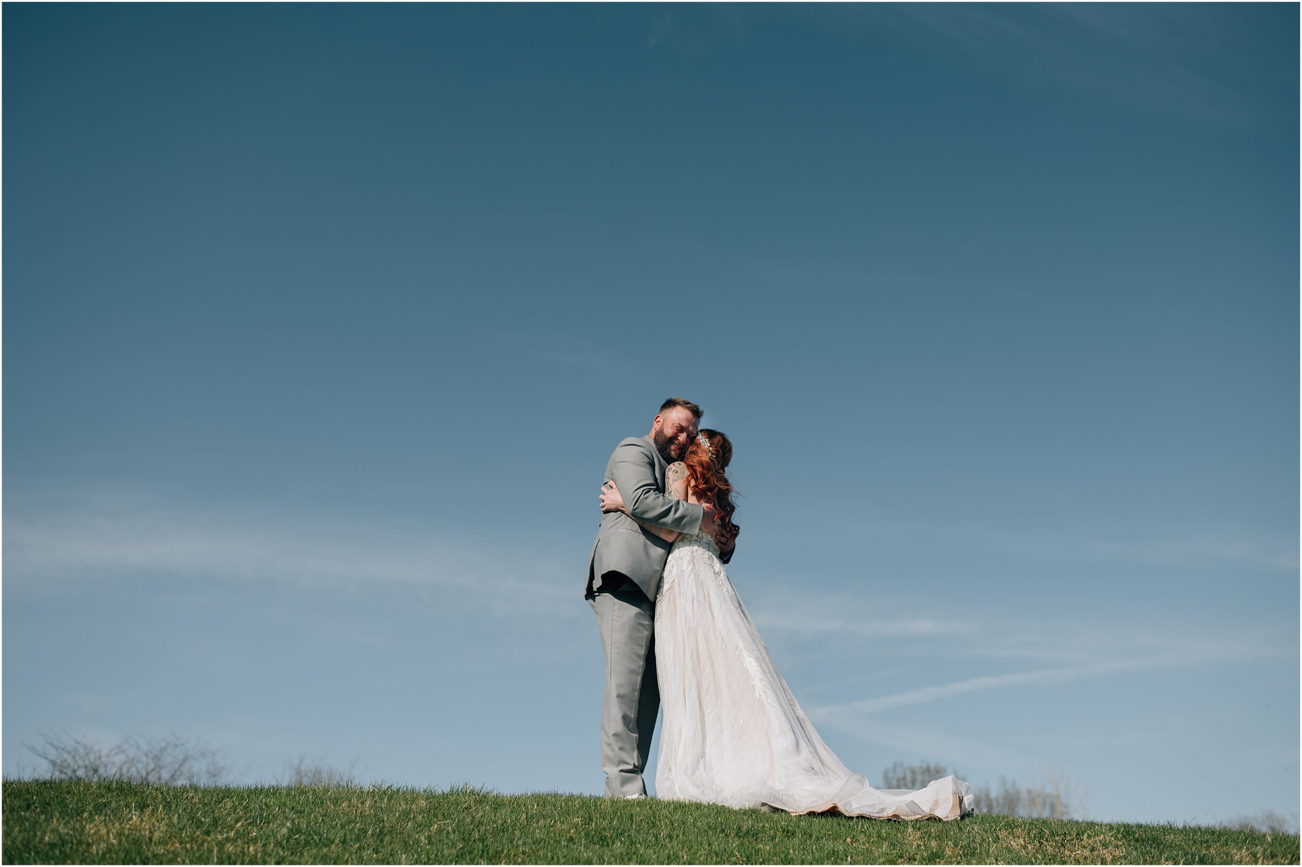 Bride in white dress with train and Groom in light grey suit share a hug on top of a hill with blue sky in the background at Honey Creek Resort on Rathbun Lake in Iowa. Photo by Anna Brace, who specializes in Omaha NE wedding photography.
