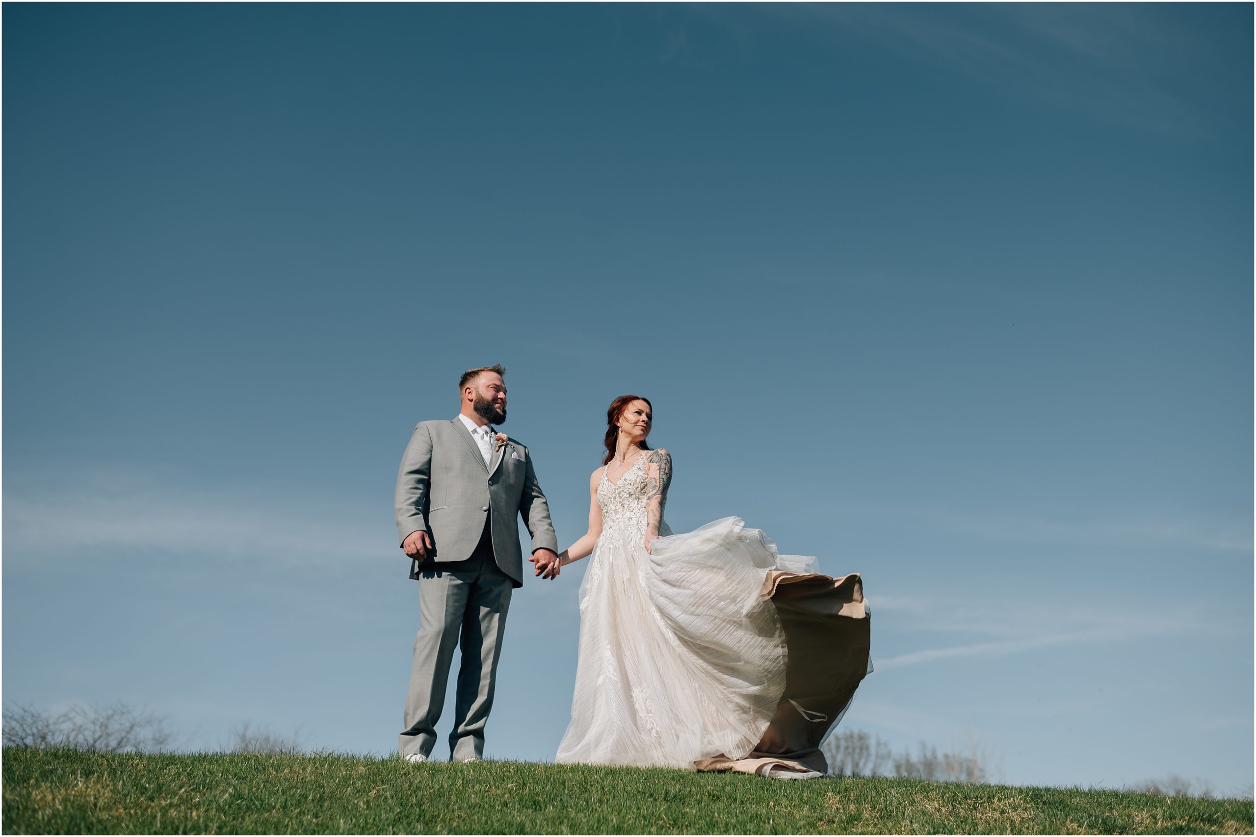 Bride in white dress with train and Groom in light grey suit walk on top of a hill with blue sky in the background at Honey Creek Resort on Rathbun Lake in Iowa. Photo by Anna Brace, who specializes in Omaha NE wedding photography.