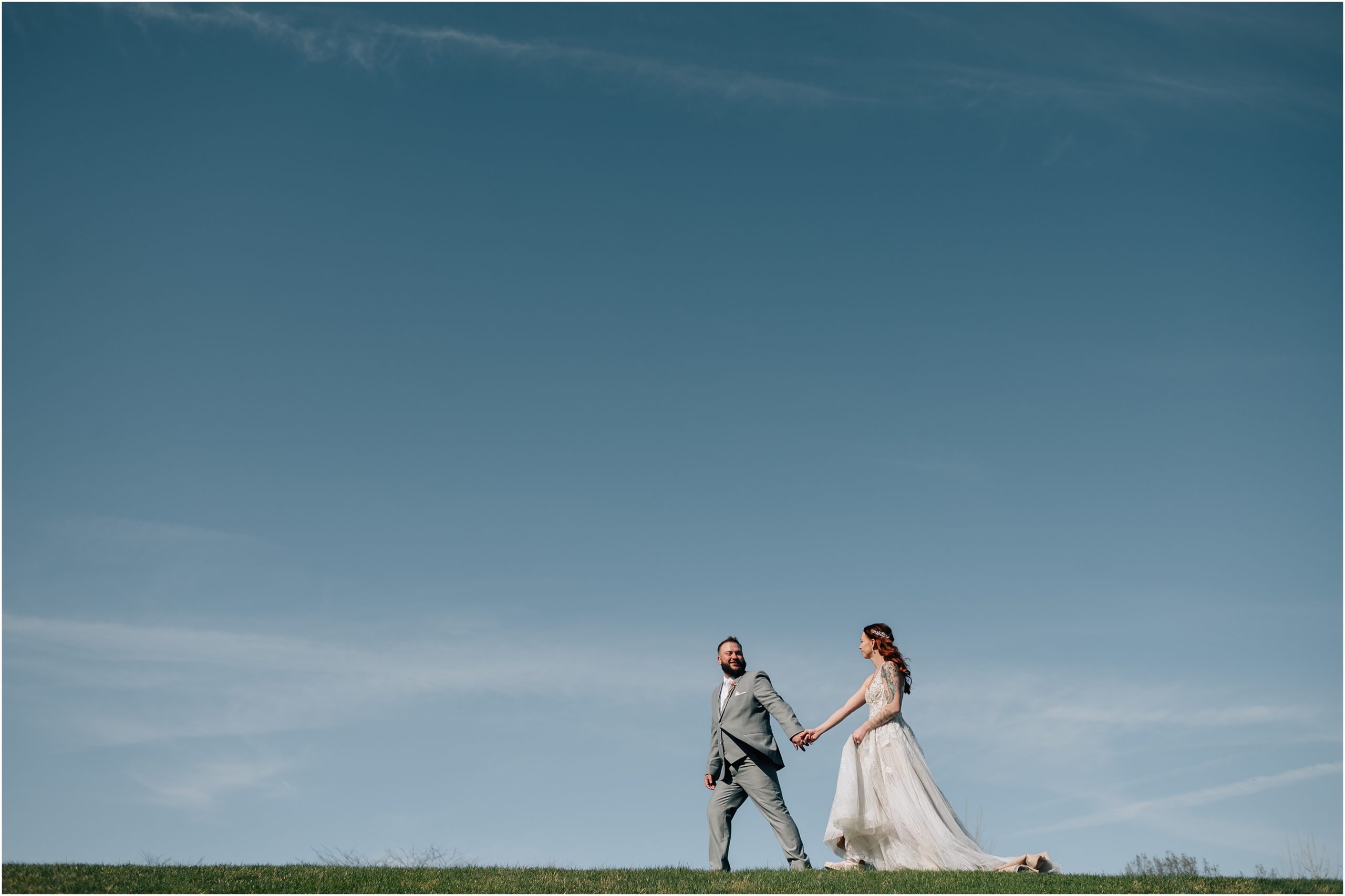 Bride in white dress with train and Groom in light grey suit walk on top of a hill with blue sky in the background at Honey Creek Resort on Rathbun Lake in Iowa. Photo by Anna Brace, who specializes in Omaha NE wedding photography.