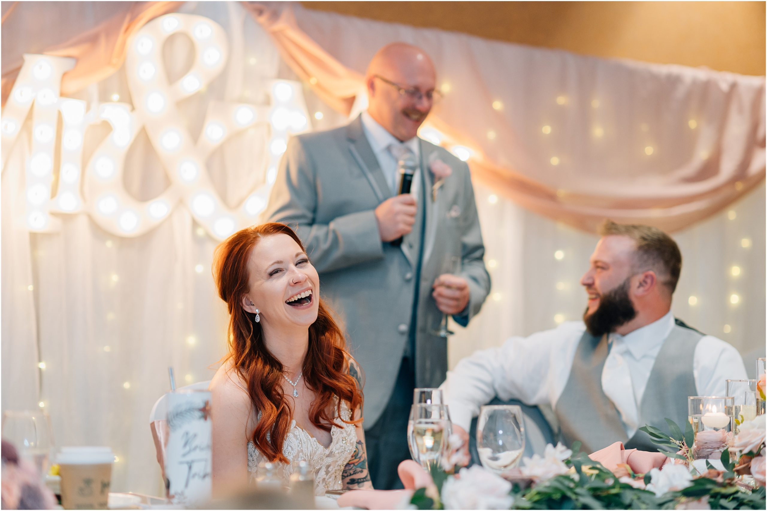 Bride laughs while her dad makes a joke during the wedding speeches at Honey Creek Resort in Iowa. By Anna Brace who specializes in Omaha Nebraska wedding photography