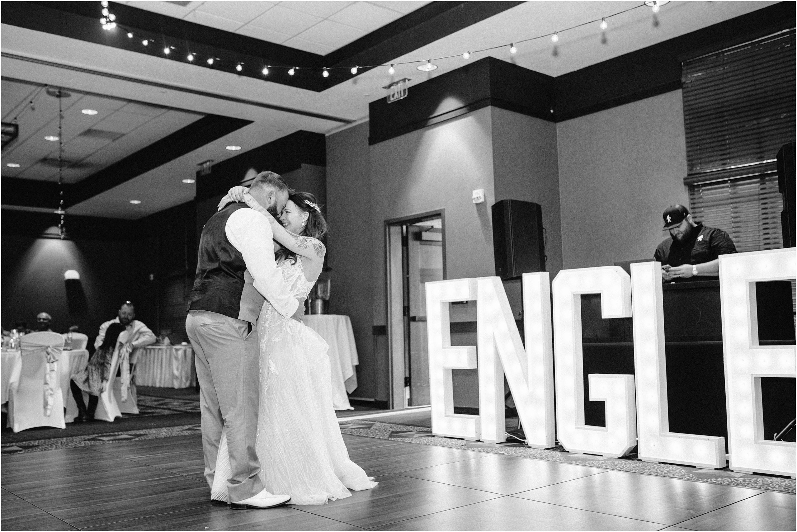 Black and white photo of bride and groom dancing with large marquee letters of their last name at Honey Creek Resort, Iowa. Photo by Omaha Nebraska Wedding Photographer, Anna Brace