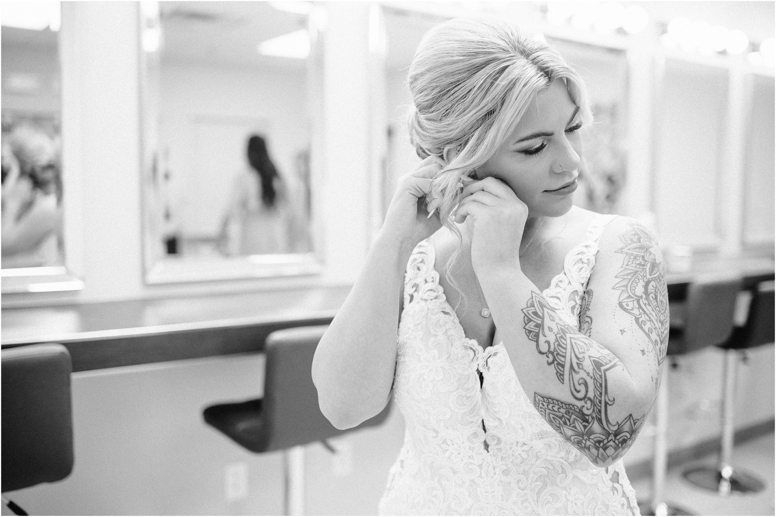 Black and white photo of bride putting on her earrings. Bride is wearing a sparkly, lace gown with a deep plunging neckline and has a sleeve of tatoos. Omaha Wedding Venue the Soiree Room taken by Anna Brace who specializes in Omaha Wedding Photography