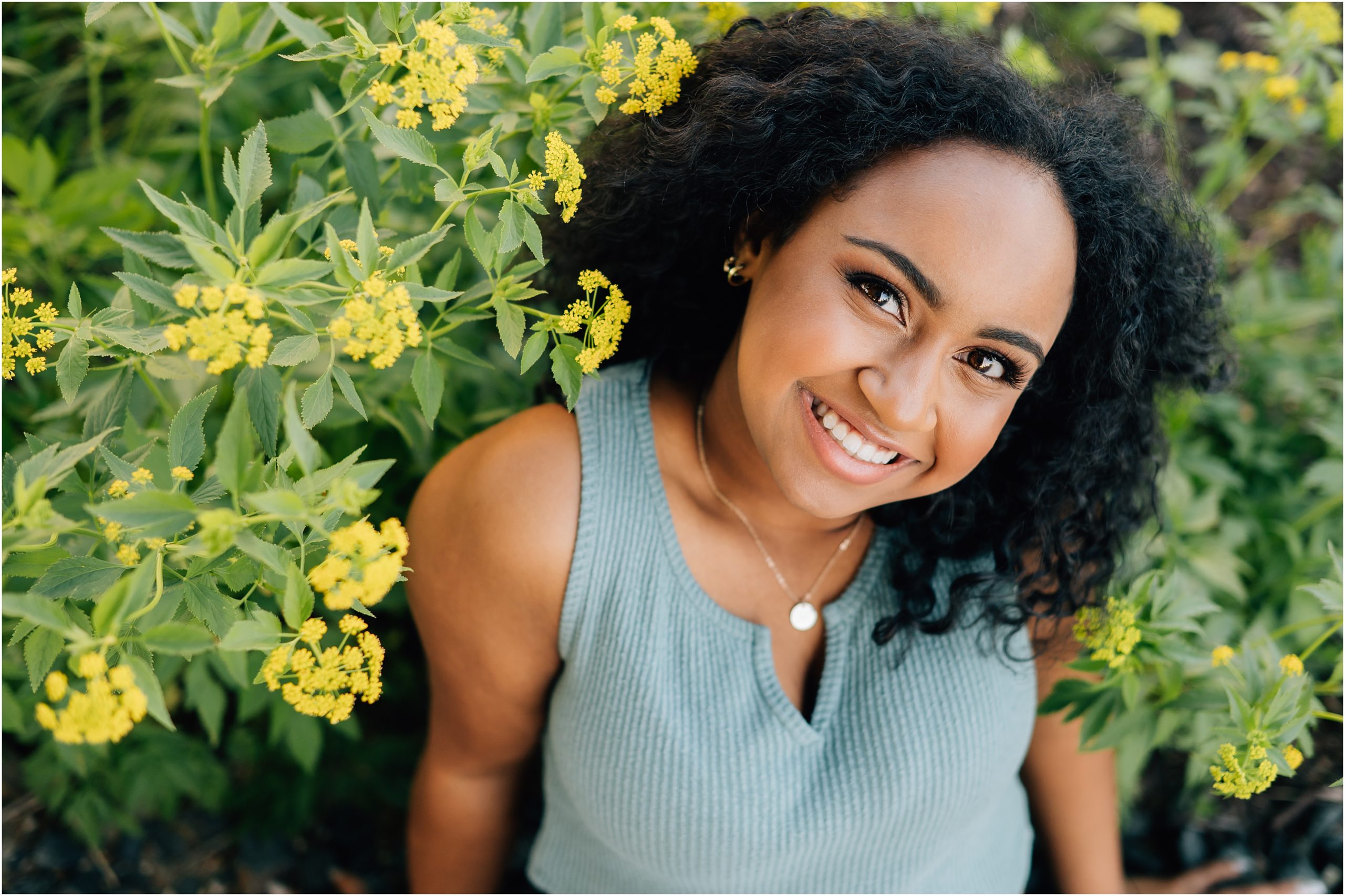 Girl with black curly hair and light blue shirt smiles up at the camera, surrounded by yellow flowers. Photo by Anna Brace who specializes in Harlan IA Photography