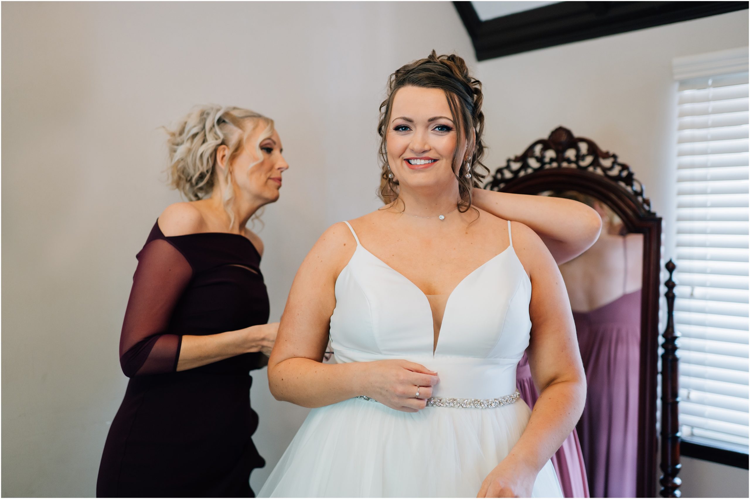 Bride smiles at the camera while her mom buttons her wedding dress at Rollins Mansion in Des Moines Iowa. Photo by Anna Brace, an omaha wedding photographer.