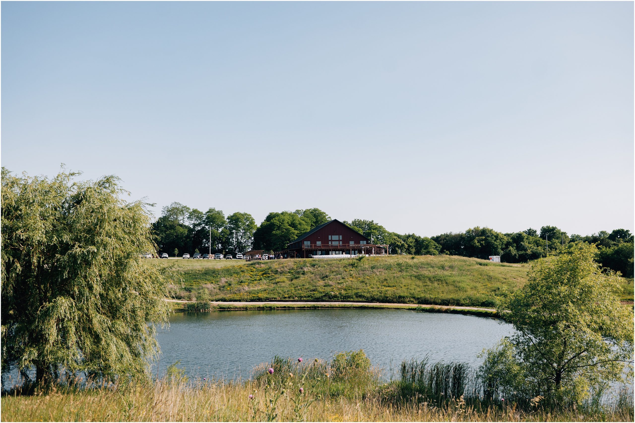 A photo of a Des Moines wedding venue, Carper Vineyard and Winery, from across the lake. The red barn is surrounded by trees and a field. Photo of the wedding venue in des moines taken by Anna Brace Photography, an Omaha Wedding Photographer. 