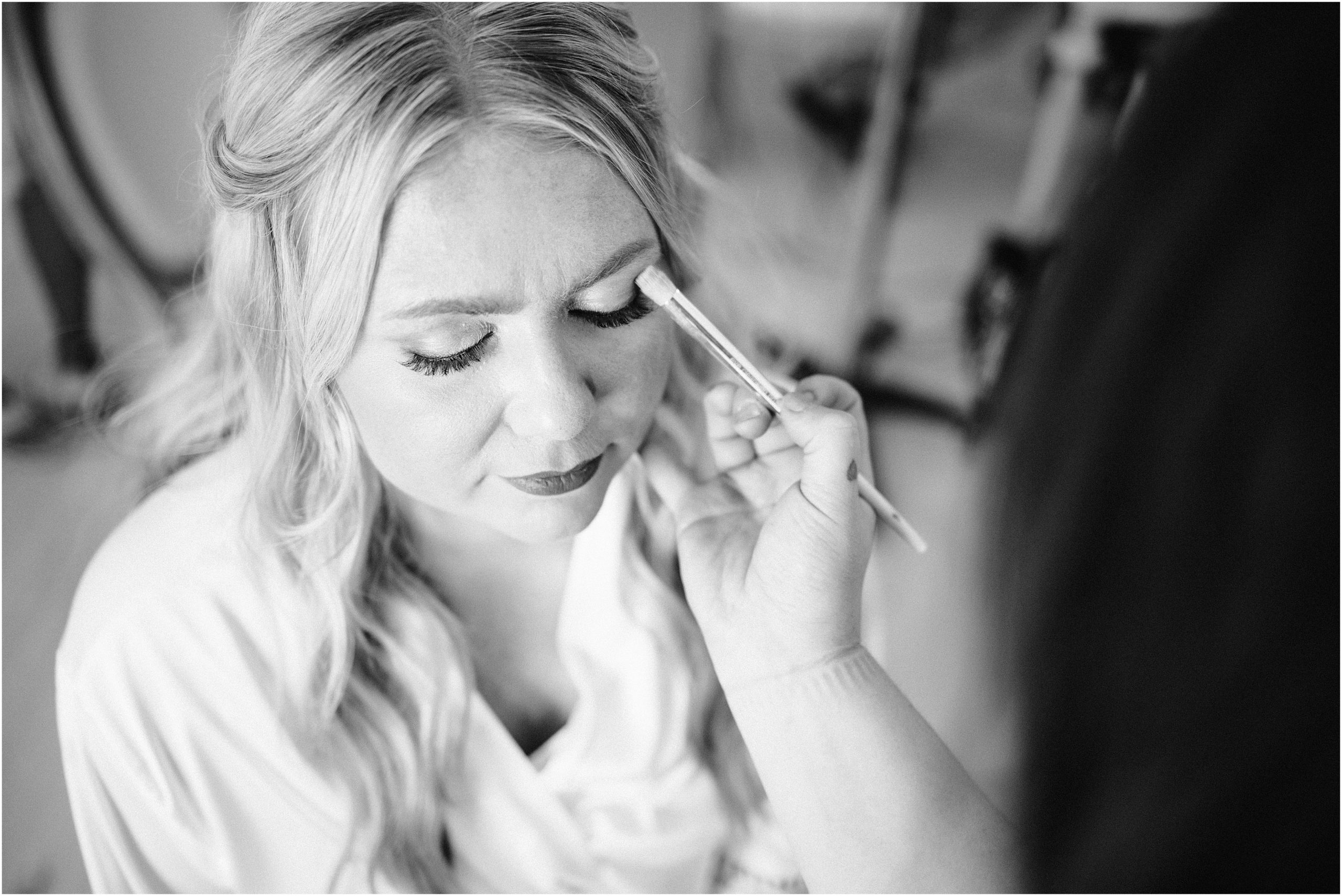 Black and white photo of bride getting her makeup done on her wedding day at Carper Vineyard, a Des Moines wedding venue. Photo by Omaha Wedding Photographer, Anna Brace.