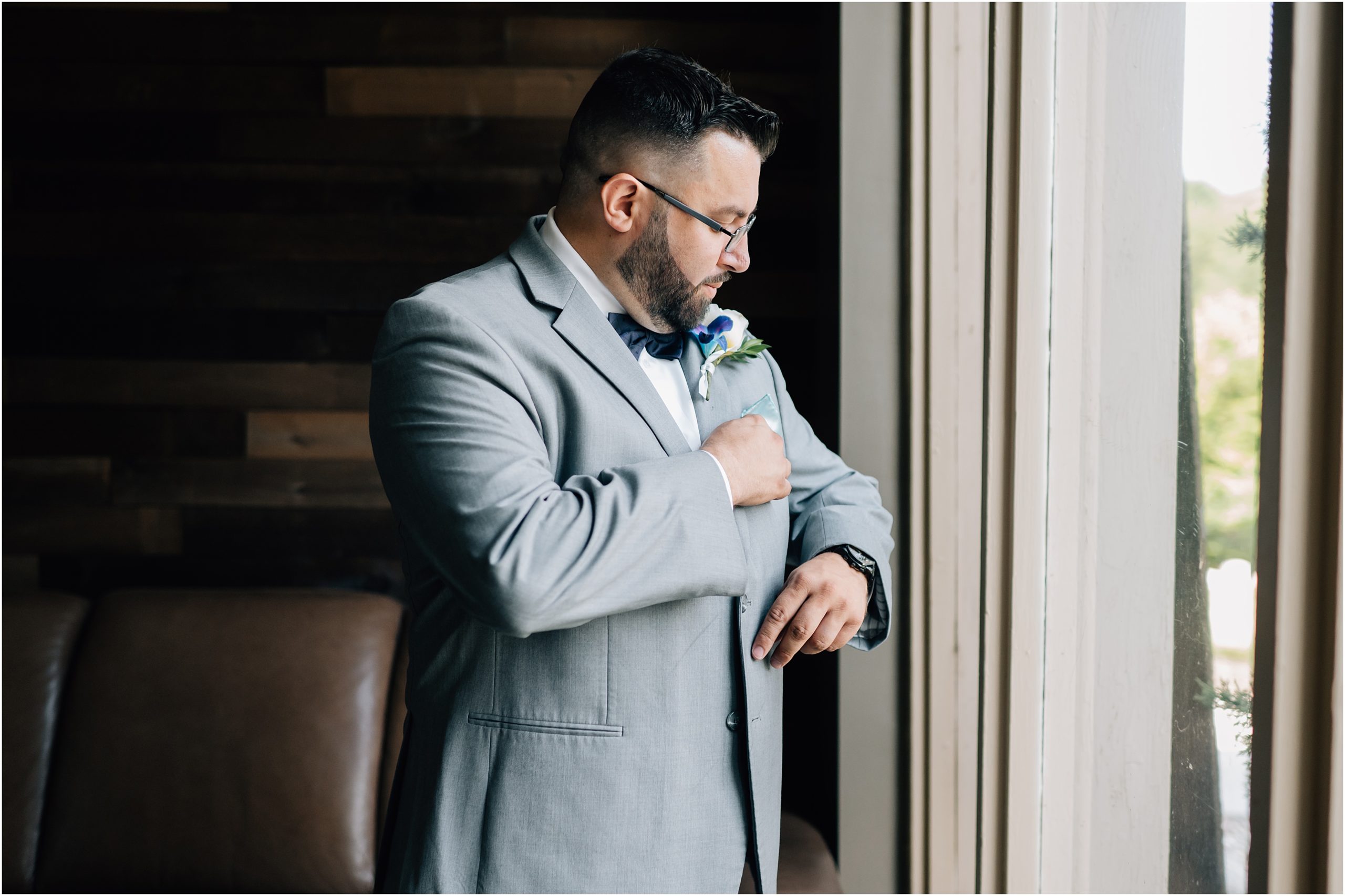 Groom in light gray suit with purple and white boutonniere adjusts his light blue pocket square while standing by a window at the omaha wedding venue A View in Fontenelle Hills. Photo was taken by Omaha Wedding Photographer, Anna Brace.