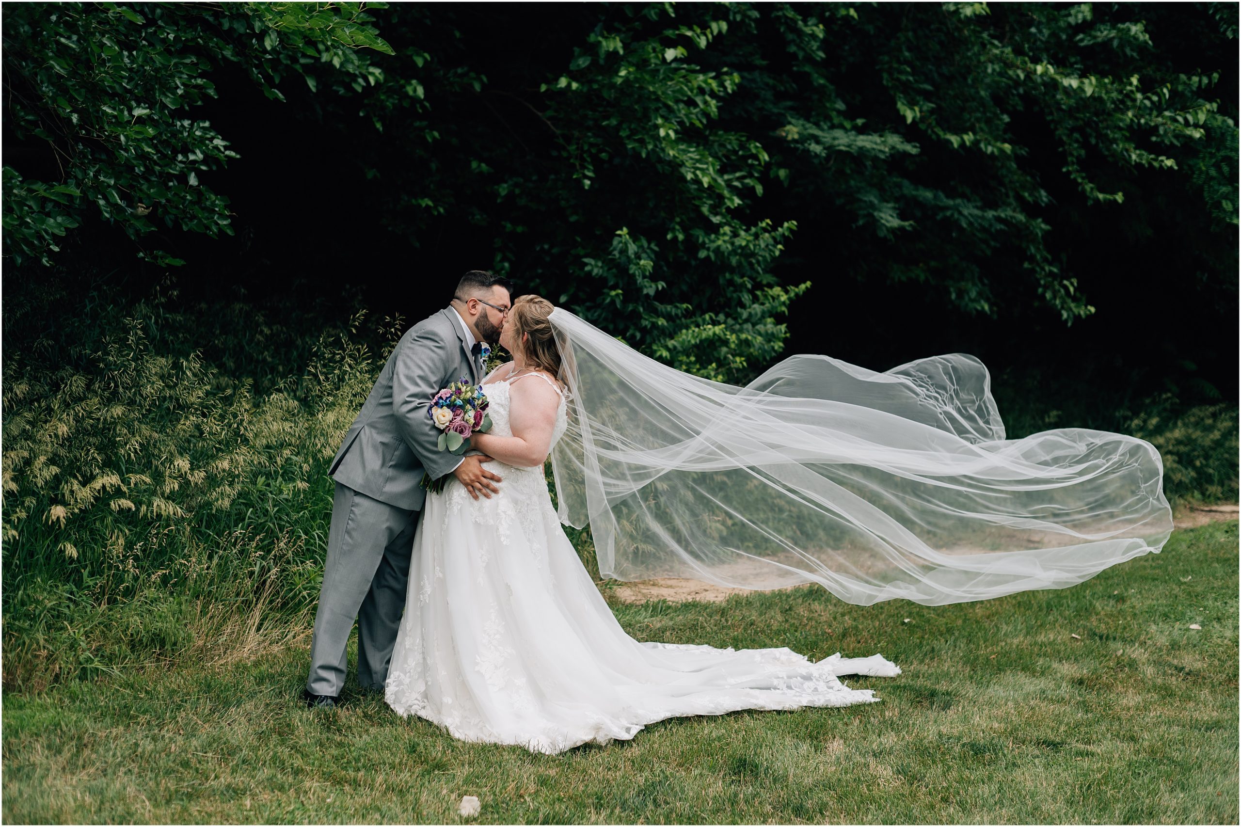 Bride, in white lace gown with cathedral length veil, and Groom, in a light grey suit, kiss while the bride's long veil blows in the wind. Photo taken at an Omaha wedding venue A View in Fontenelle Hills by Omaha wedding photographer Anna Brace.