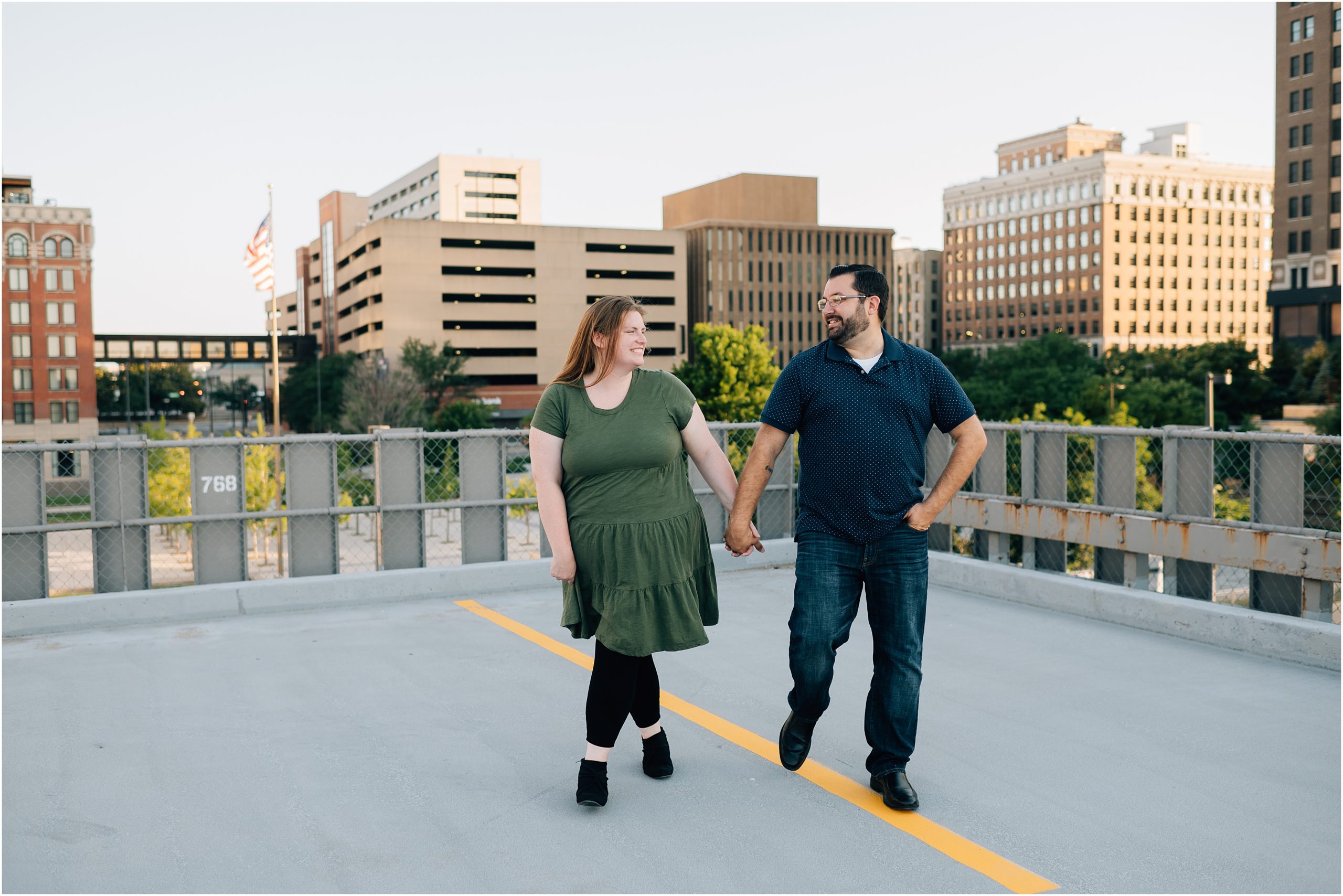 Woman wearing green dress and man wearing blue shirt and jeans walk hand in hand on top of a parking garage in downtown Omaha. Photo taken by Anna Brace, an Omaha wedding photographer. 
