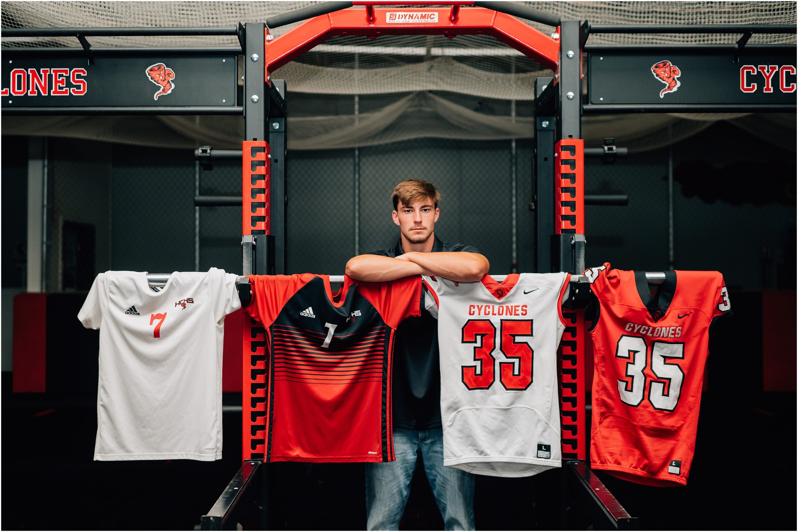 Weight Room senior picture ideas - photograph by Anna Brace, a Harlan Iowa Portrait Photographer of a boy standing underneath a weight lifting set with his jersey's strung along the weight bar.