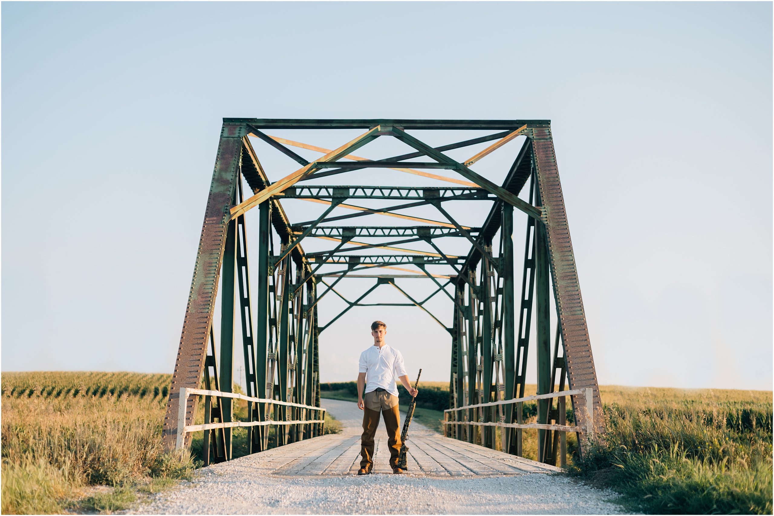 Photograph of an old green/brown bridge with a senior boy holding a gun in one hand. Boy is wearing white and brown. Photograph by Harlan Iowa photographer, Anna Brace.