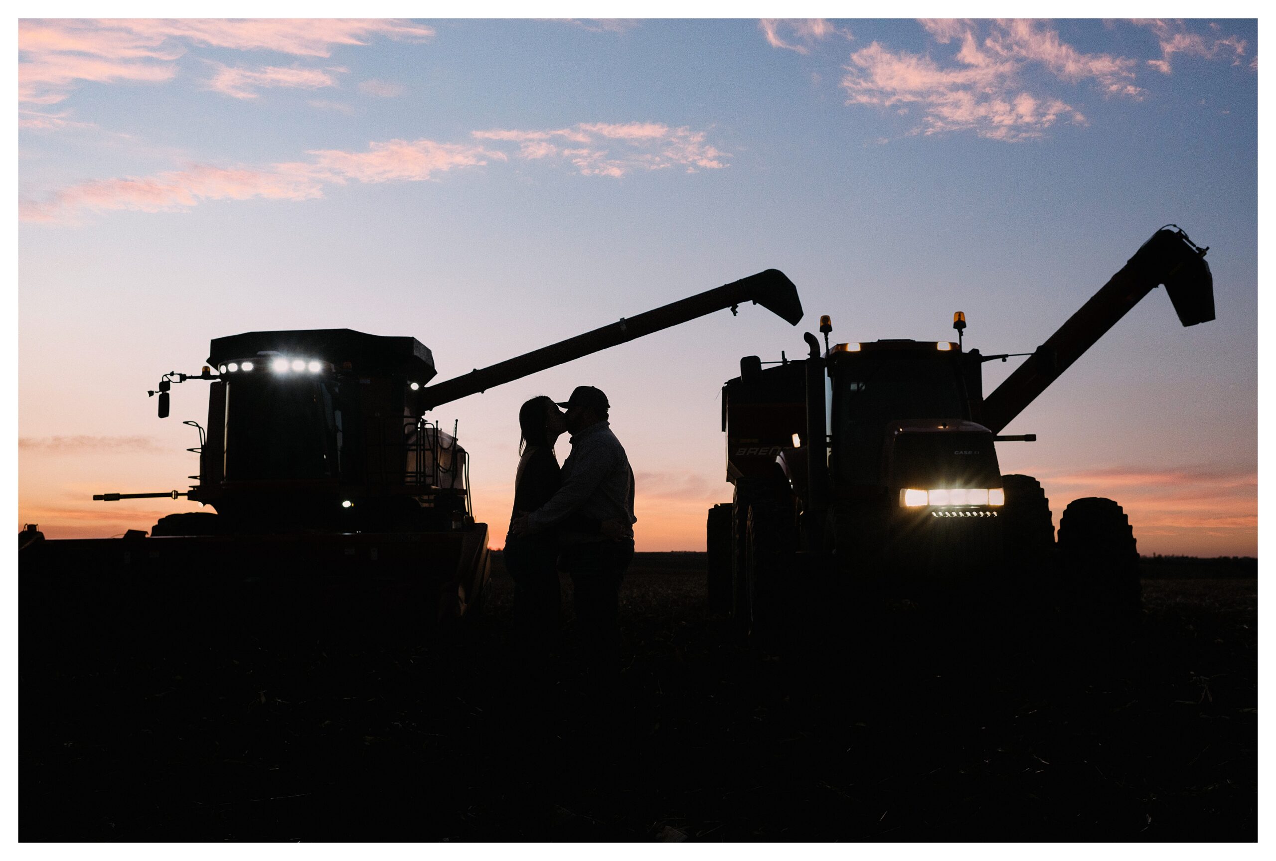 Silhouette of an engaged couple kissing in from of a combine and a tractor pulling an auger wagon. The sky has a gorgeous sunset behind them. Photo by Anna Brace, an Omaha NE Wedding Photographer.