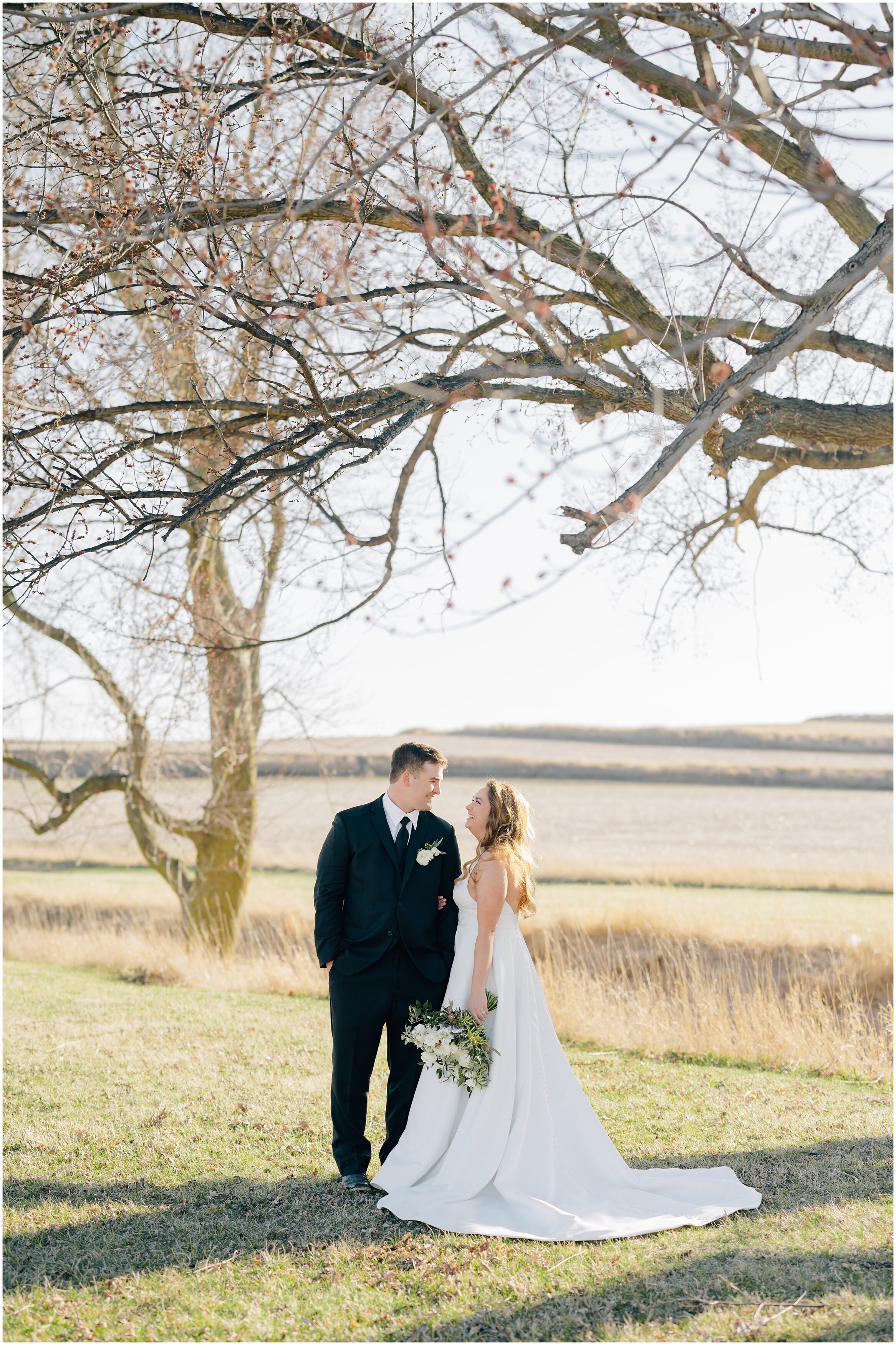 Bride white gown with a greenery  white rose bouquet smiles up at groom, wearing a black suit and cowboy boots during their Palace Event Center wedding in Treynor Ia. Photo by Anna brace, a wedding photographer in Omaha.