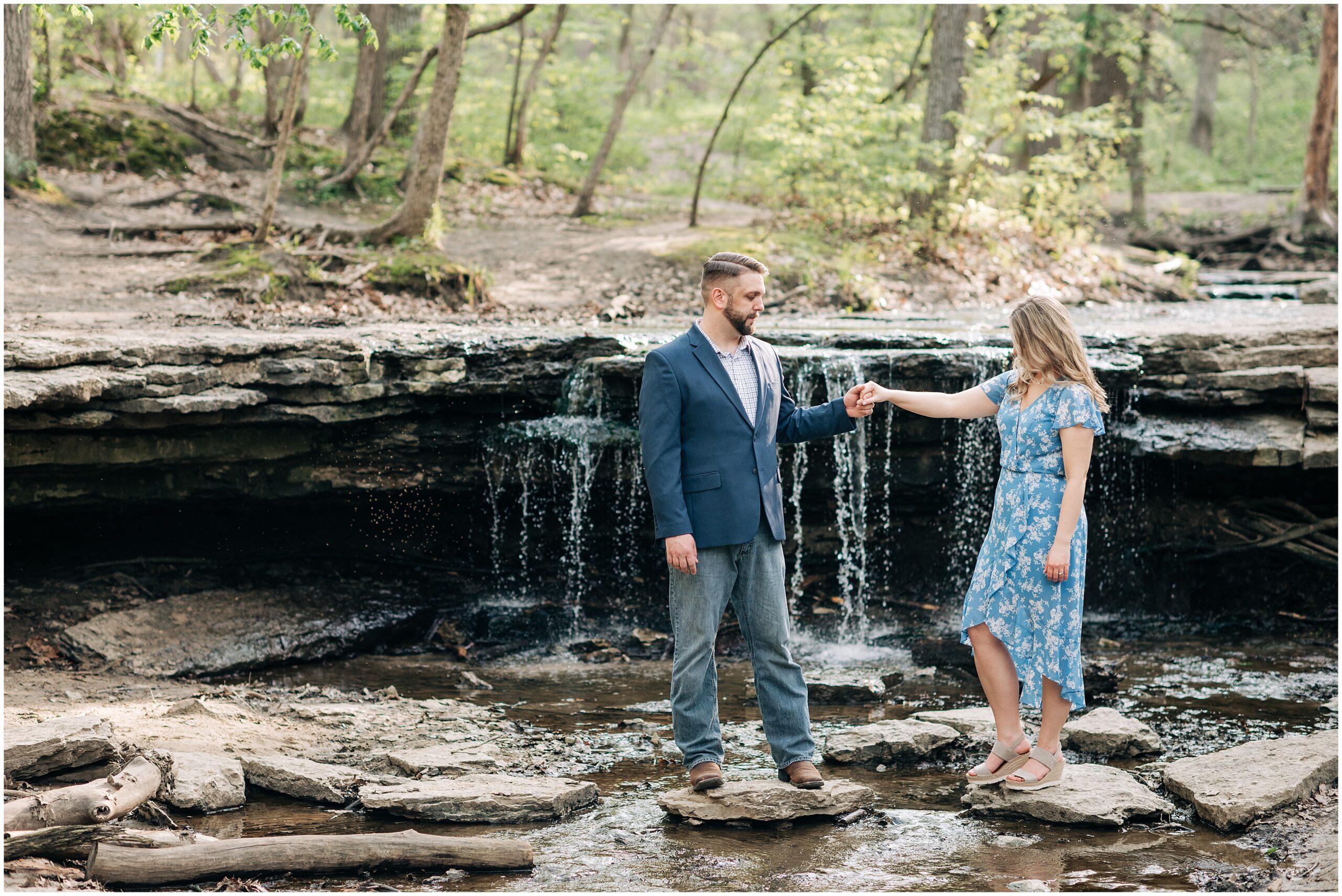 Girl in blue floral dress and guy in suit jacket and jeans stand on rocks in front of the waterfall at Platte River State Park during their engagement pictures. Photo by Anna Brace, a Nebraska wedding photographer.