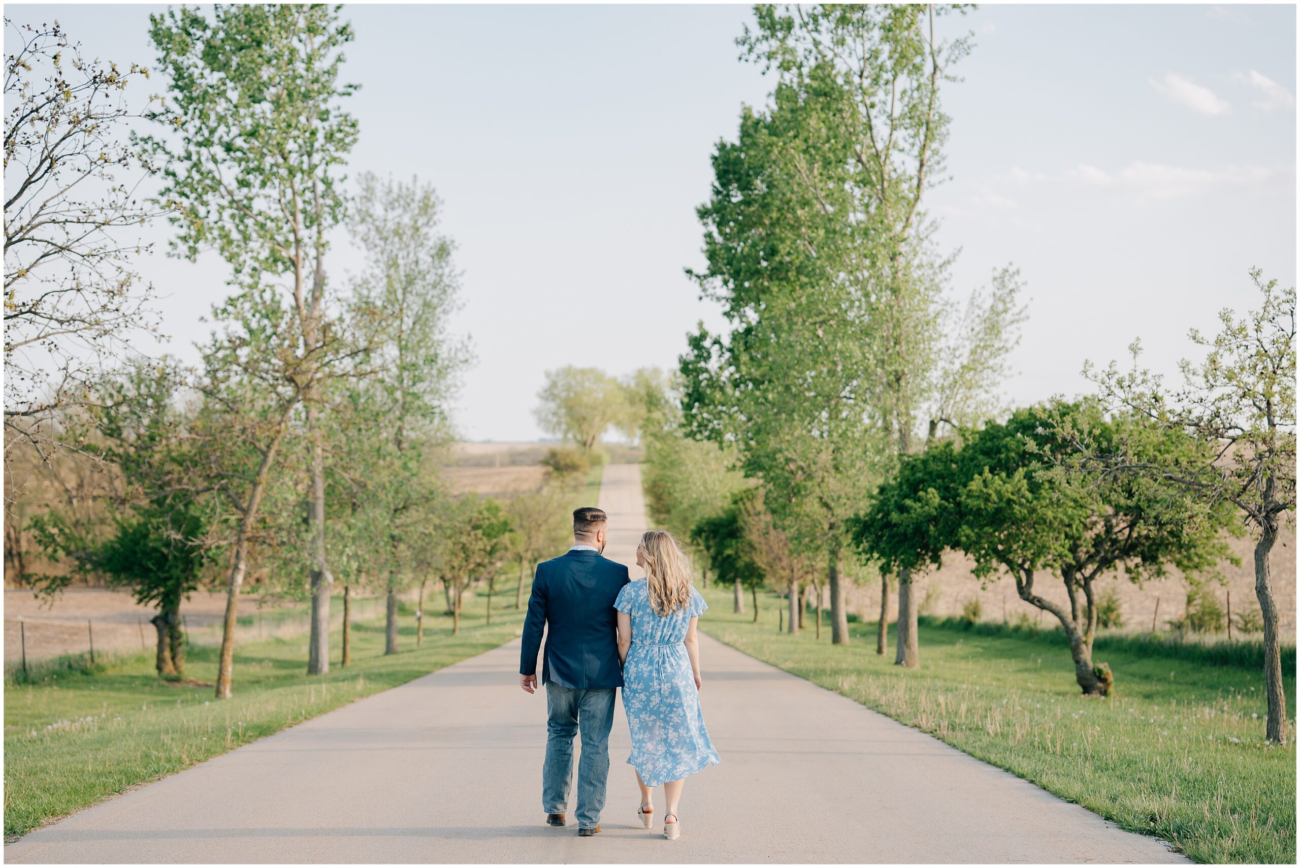 Girl wearing a blue floral dress and guy wearing a blue suit jacket and jeans walk down a road lined with trees. Photo by Omaha ne wedding photographer, Anna Brace.